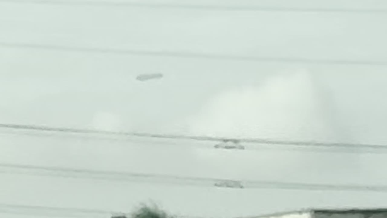 Enlarged frame: a dark, level, elongated object against pale cloud, above the power lines.